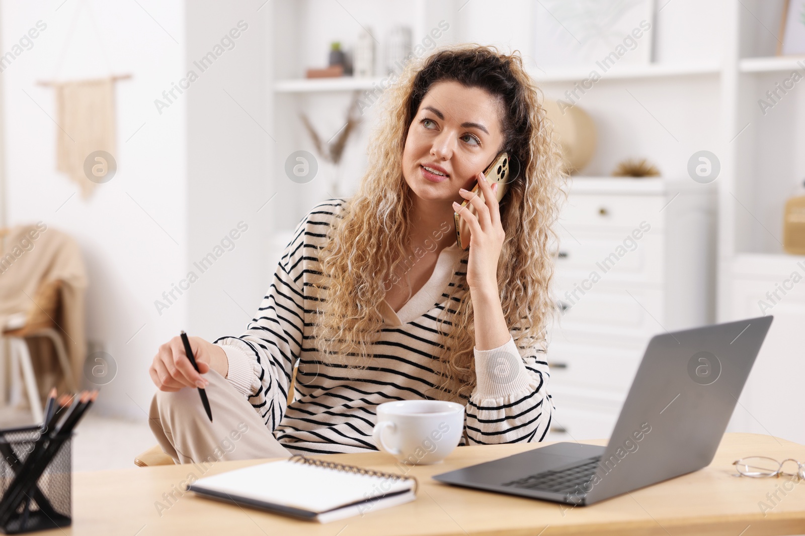 Smiling woman talking by smartphone at desk with laptop in home office Photo of Smiling woman talking by smartphone at desk with laptop in home office