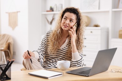 Photo of Smiling woman talking by smartphone at desk with laptop in home office