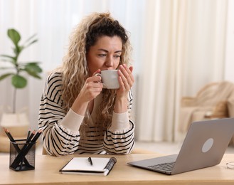 Photo of Woman with cup of hot drink working on laptop at desk in home office