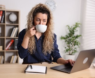 Photo of Woman with cup of hot drink working on laptop at desk in home office
