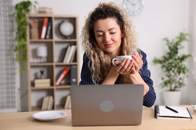 Photo of Woman with cup of hot drink working on laptop at desk in home office