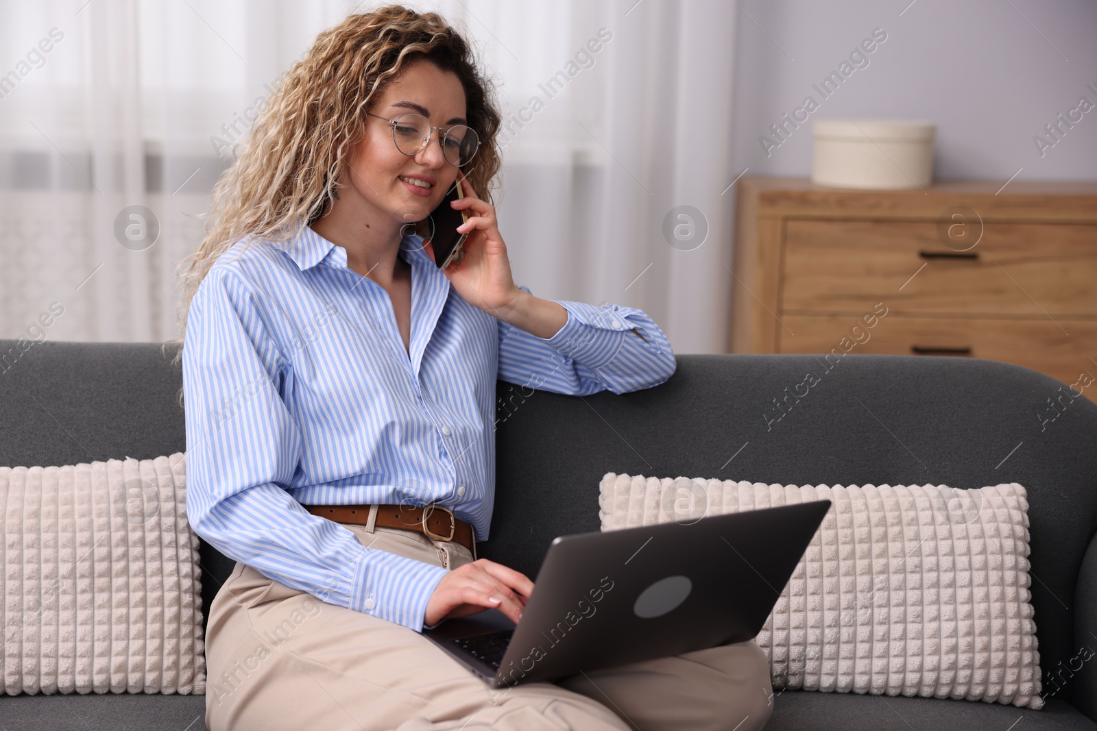 Smiling woman with laptop talking by smartphone on sofa in home office Photo of Smiling woman with laptop talking by smartphone on sofa in home office