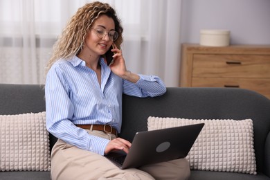 Photo of Smiling woman with laptop talking by smartphone on sofa in home office