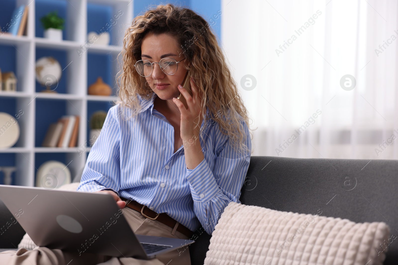 Woman with laptop talking by smartphone on sofa in home office Photo of Woman with laptop talking by smartphone on sofa in home office