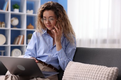 Photo of Woman with laptop talking by smartphone on sofa in home office