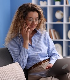 Photo of Smiling woman with laptop talking by smartphone on sofa in home office