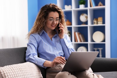 Photo of Smiling woman with laptop talking by smartphone on sofa in home office