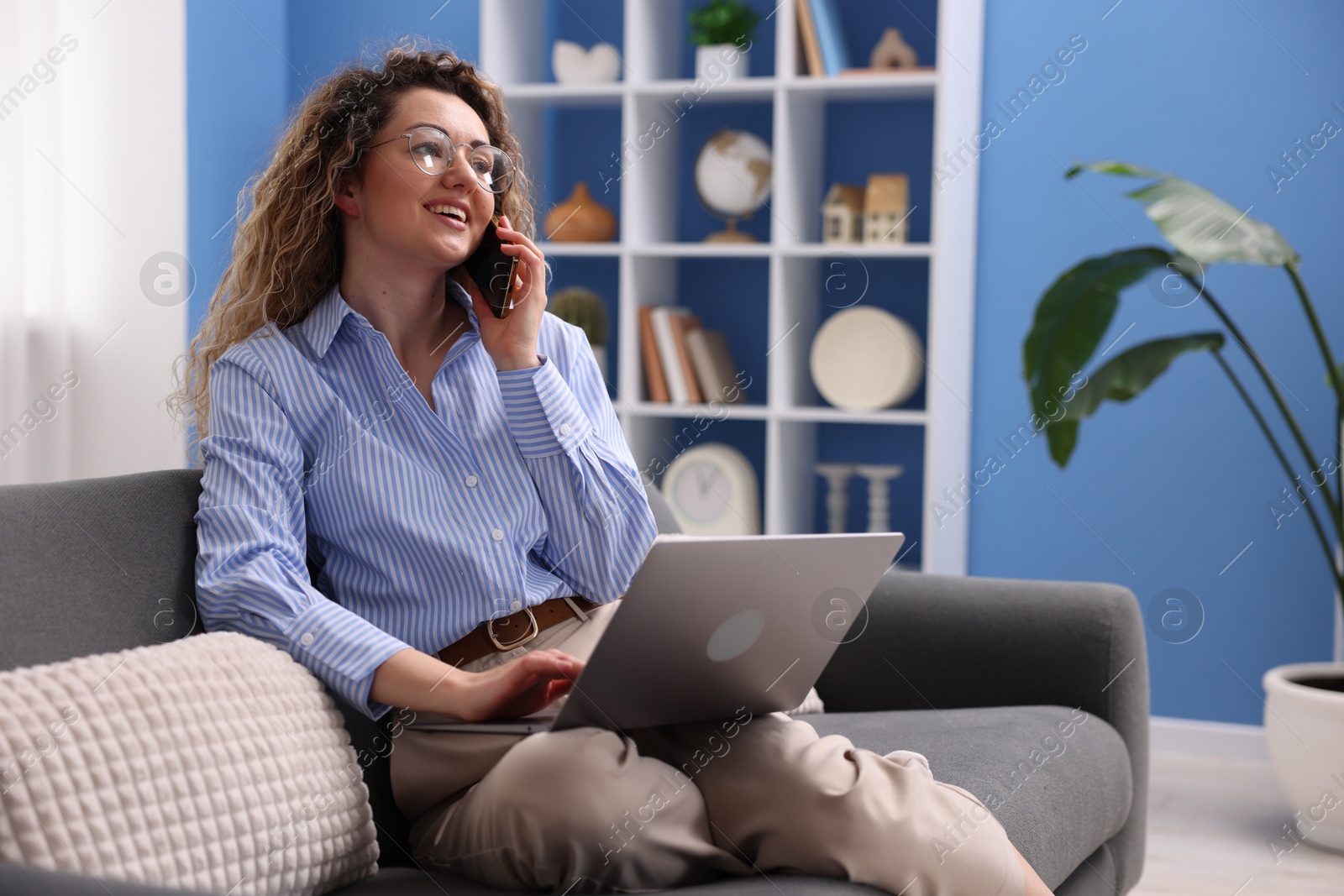 Smiling woman with laptop talking by smartphone on sofa in home office Photo of Smiling woman with laptop talking by smartphone on sofa in home office