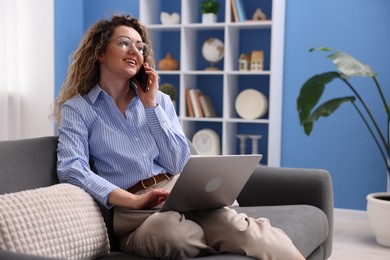 Photo of Smiling woman with laptop talking by smartphone on sofa in home office