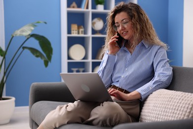 Photo of Smiling woman with laptop talking by smartphone on sofa in home office