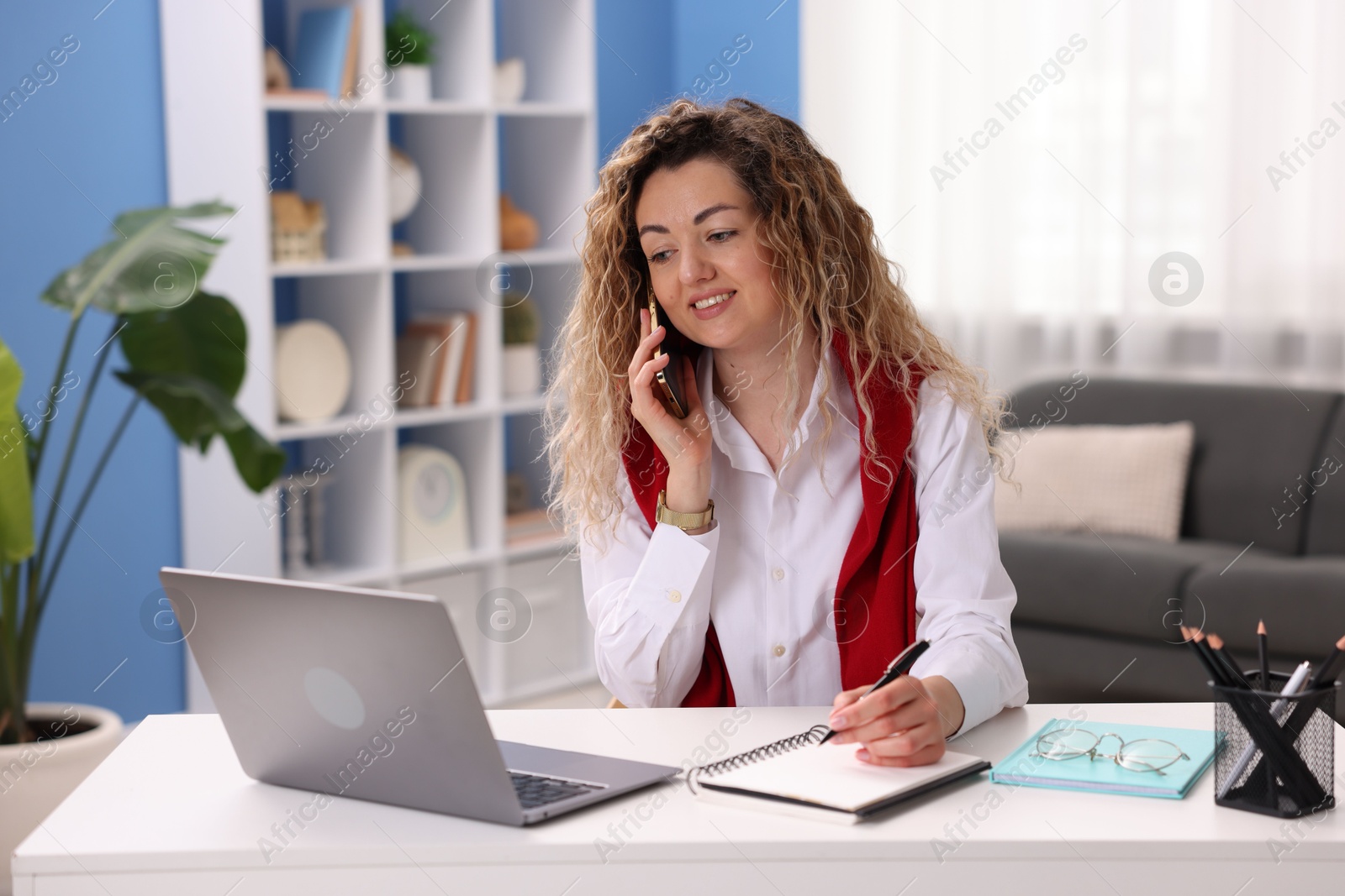 Smiling woman with laptop talking by smartphone at desk in home office Photo of Smiling woman with laptop talking by smartphone at desk in home office