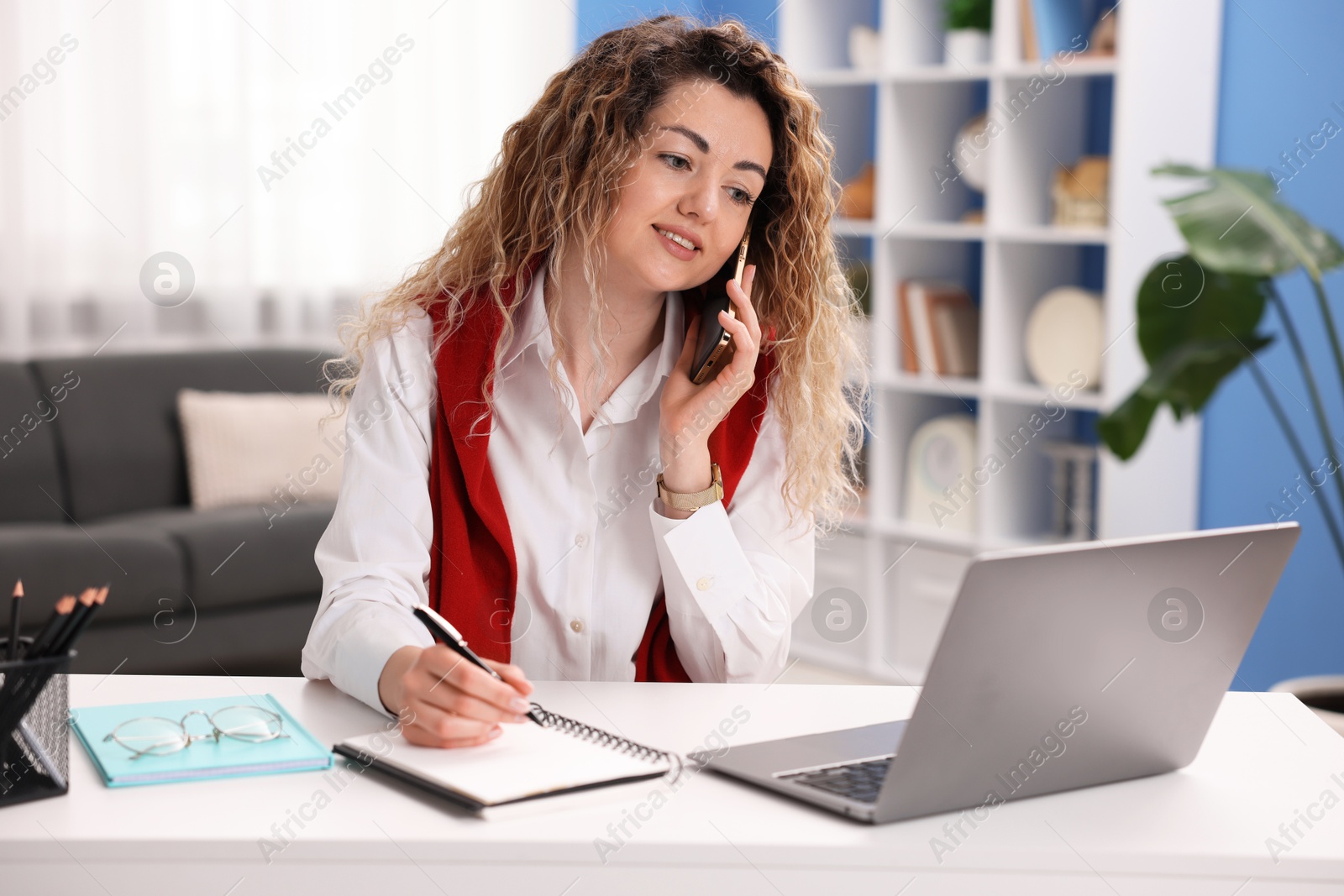 Smiling woman with laptop talking by smartphone at desk in home office Photo of Smiling woman with laptop talking by smartphone at desk in home office