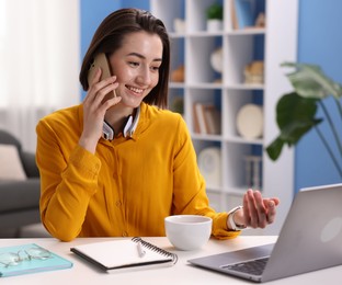 Photo of Smiling woman with laptop talking by smartphone at desk in home office