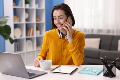 Photo of Smiling woman with laptop talking by smartphone at desk in home office