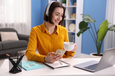 Photo of Smiling woman with smartphone and cup of hot drink at desk in home office