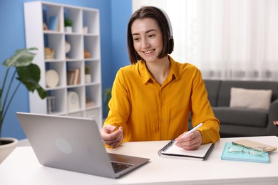 Photo of Smiling woman in headphones having videochat by laptop at desk in home office
