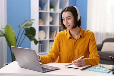 Photo of Woman in headphones having videochat by laptop at desk in home office