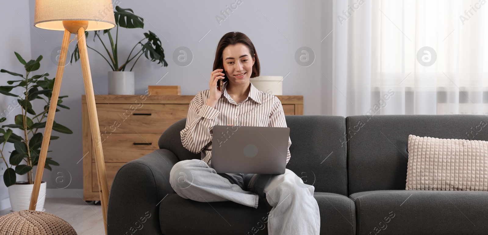 Smiling woman with laptop talking by smartphone on sofa in home office Photo of Smiling woman with laptop talking by smartphone on sofa in home office