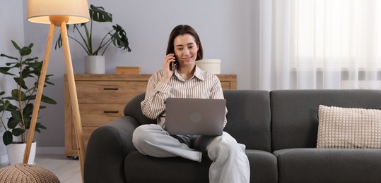 Photo of Smiling woman with laptop talking by smartphone on sofa in home office