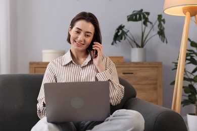 Photo of Smiling woman with laptop talking by smartphone on sofa in home office