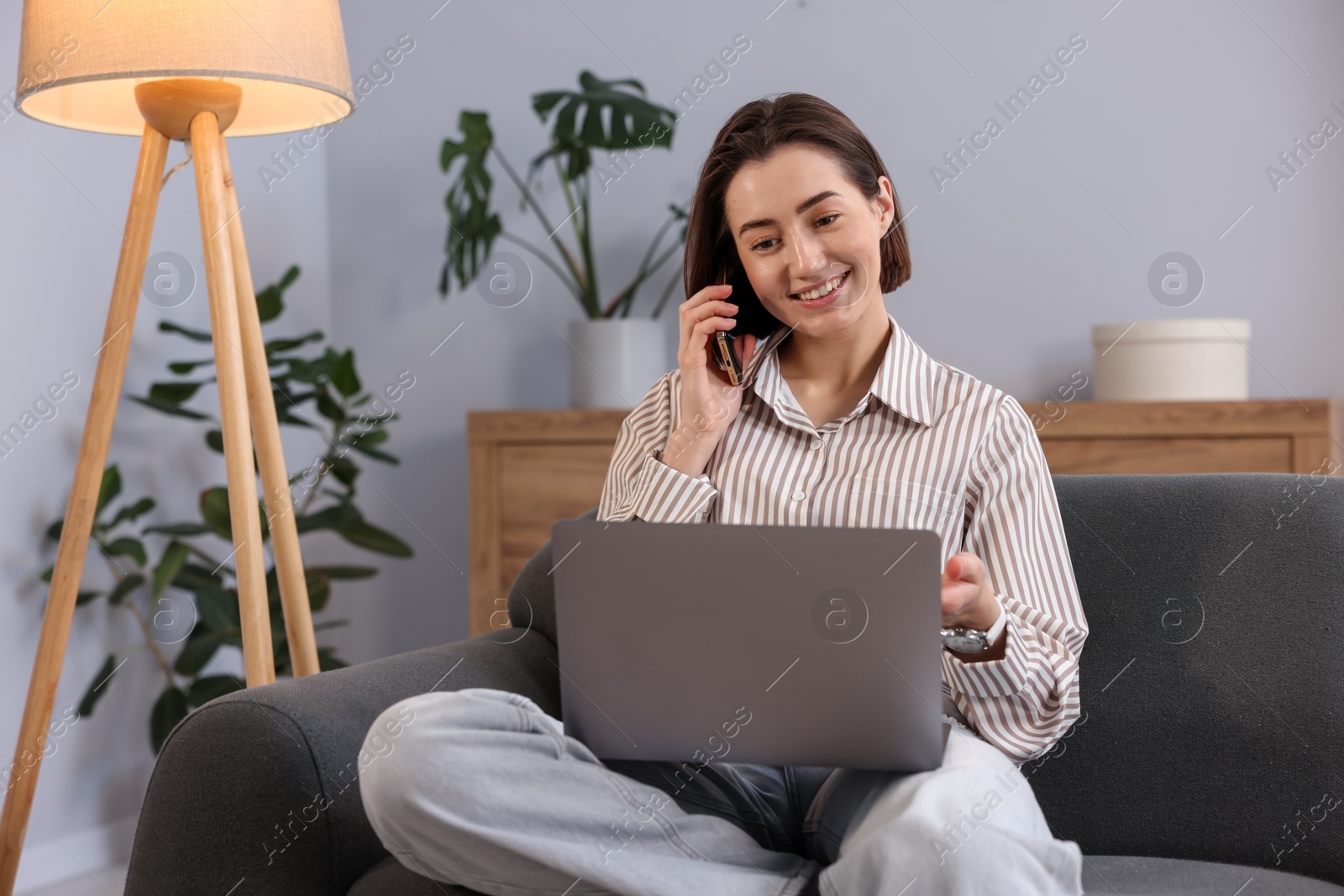 Smiling woman with laptop talking by smartphone on sofa in home office Photo of Smiling woman with laptop talking by smartphone on sofa in home office