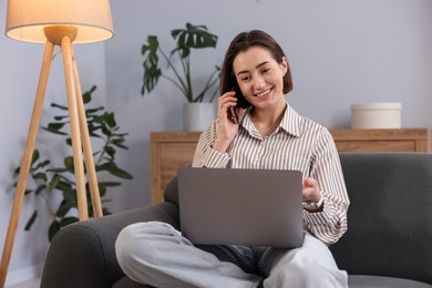 Photo of Smiling woman with laptop talking by smartphone on sofa in home office
