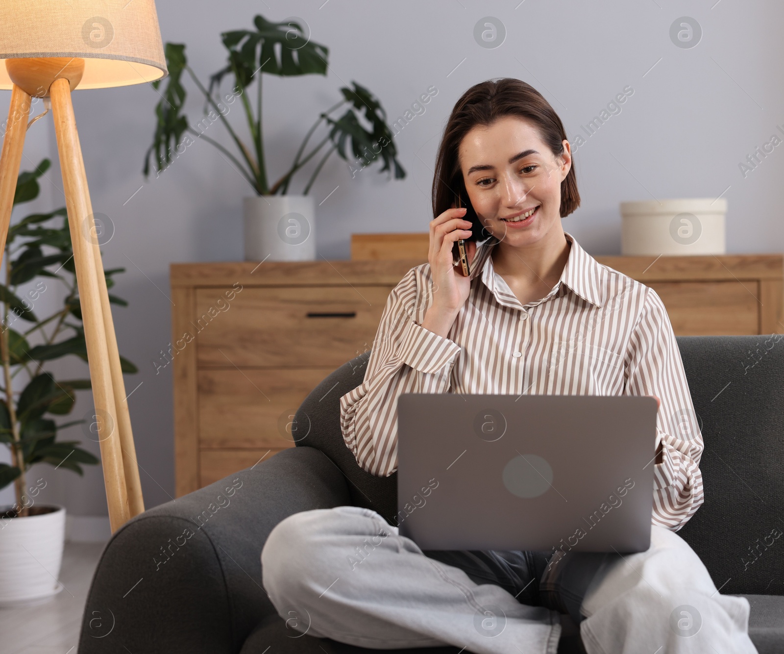 Smiling woman with laptop talking by smartphone on sofa in home office Photo of Smiling woman with laptop talking by smartphone on sofa in home office