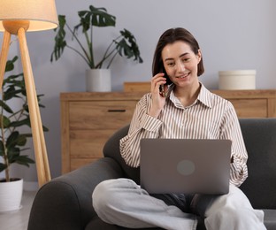 Photo of Smiling woman with laptop talking by smartphone on sofa in home office