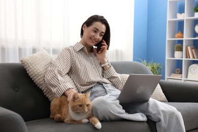 Photo of Smiling woman talking by smartphone with laptop and cat on sofa in home office