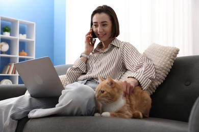Photo of Smiling woman talking by smartphone with laptop and cat on sofa in home office, low angle view