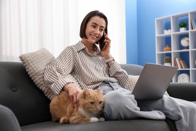 Photo of Smiling woman talking by smartphone with laptop and cat on sofa in home office, low angle view