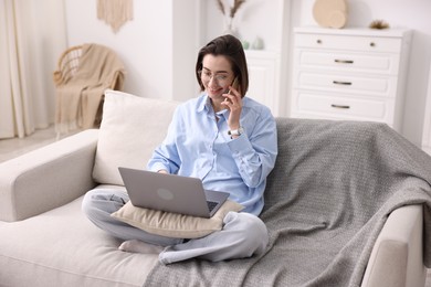 Photo of Smiling woman talking by smartphone with laptop on sofa in home office