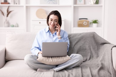 Photo of Smiling woman talking by smartphone with laptop on sofa in home office
