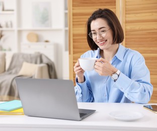 Photo of Smiling woman with cup of hot drink having videochat by laptop at desk in home office