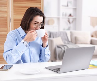 Photo of Smiling woman with cup of hot drink having videochat by laptop at desk in home office