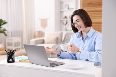 Photo of Smiling woman with cup of hot drink having videochat by laptop at desk in home office
