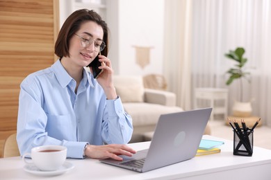 Photo of Smiling woman talking by smartphone with laptop at desk in home office