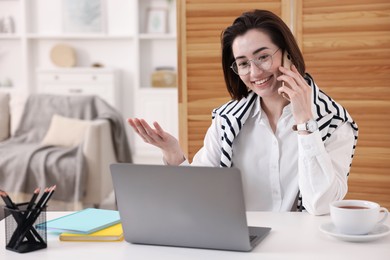 Photo of Smiling woman talking by smartphone with laptop at desk in home office