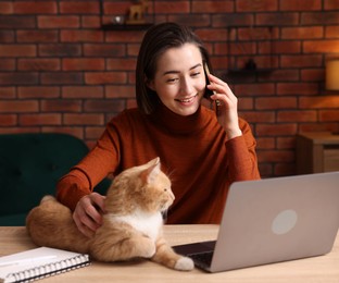 Photo of Smiling woman talking by smartphone with laptop and cat at desk in home office