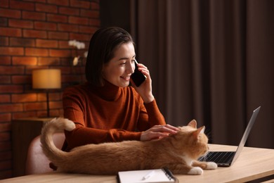 Photo of Smiling woman talking by smartphone and stroking cat at desk in home office