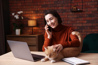 Photo of Smiling woman talking by smartphone and stroking cat at desk in home office