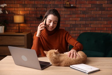 Photo of Smiling woman talking by smartphone and stroking cat at desk in home office