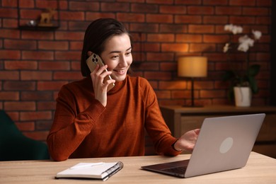 Photo of Smiling woman talking by smartphone at desk with laptop in home office