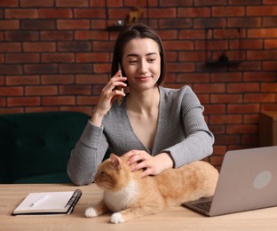 Photo of Woman talking by smartphone and stroking cat at desk in home office