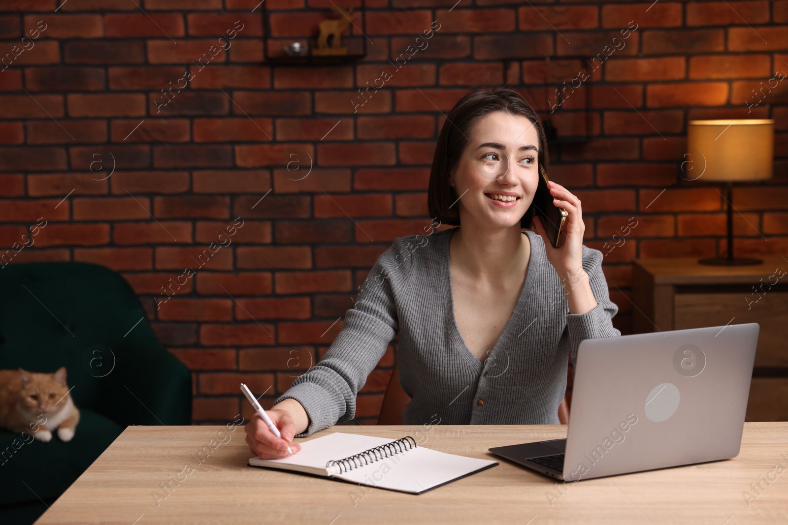 Smiling woman talking by smartphone at desk with laptop in home office Photo of Smiling woman talking by smartphone at desk with laptop in home office