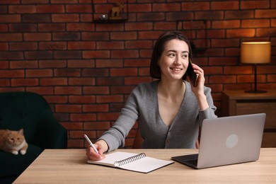 Photo of Smiling woman talking by smartphone at desk with laptop in home office