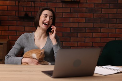 Photo of Happy woman with cat talking by smartphone at desk with laptop in home office