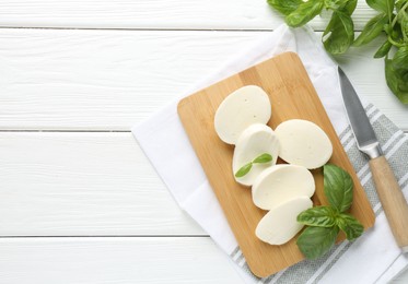 Photo of Pieces of delicious mozzarella cheese, basil leaves and knife on white wooden table, flat lay. Space for text