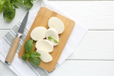 Photo of Pieces of delicious mozzarella cheese, basil leaves and knife on white wooden table, flat lay. Space for text