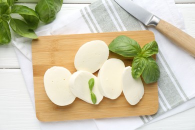Photo of Pieces of delicious mozzarella cheese, basil leaves and knife on white wooden table, flat lay